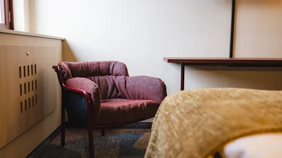 A red cushioned chair sits against a light-colored wall near a window, beside a cabinet, with a bed and table nearby.