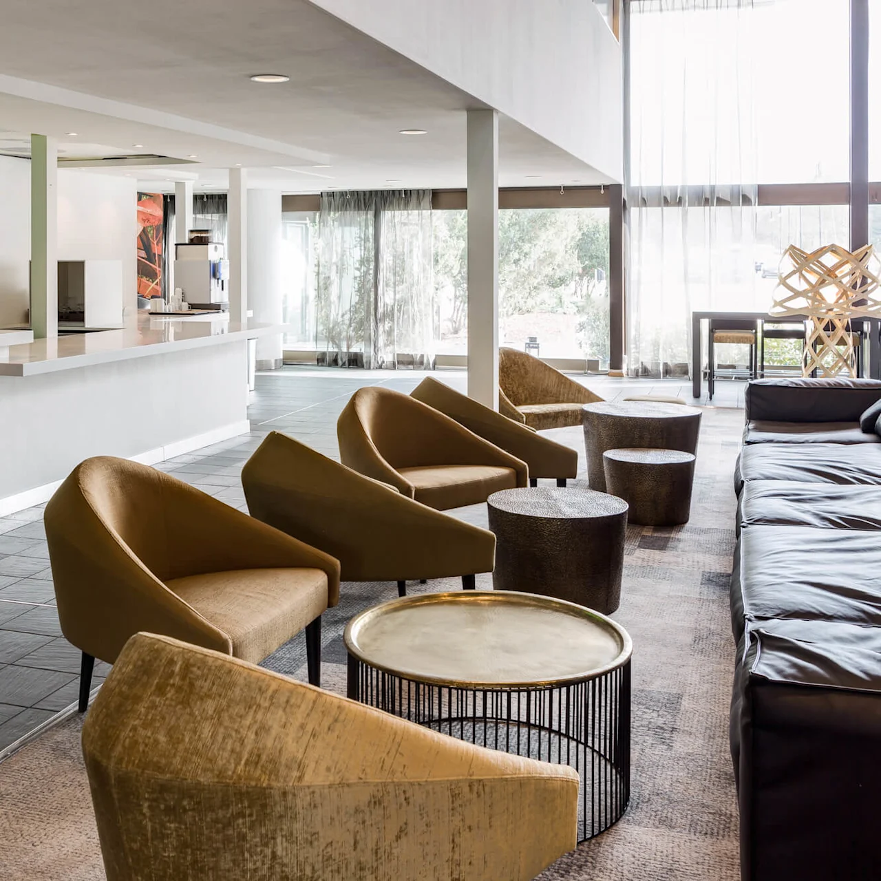 A modern lounge area features brown chairs and small round tables, set near a long counter bar with green-lit shelves, illuminated by natural light from large windows.