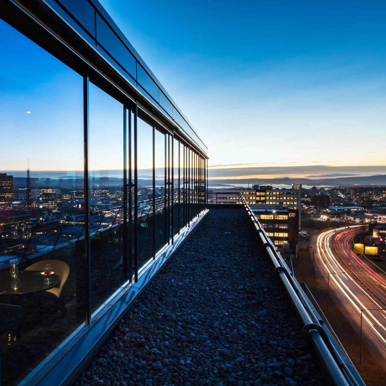Glass-walled rooftop restaurant reflects the night city skyline, with interior tables dimly lit. Adjacent road below shows light trails from moving vehicles.