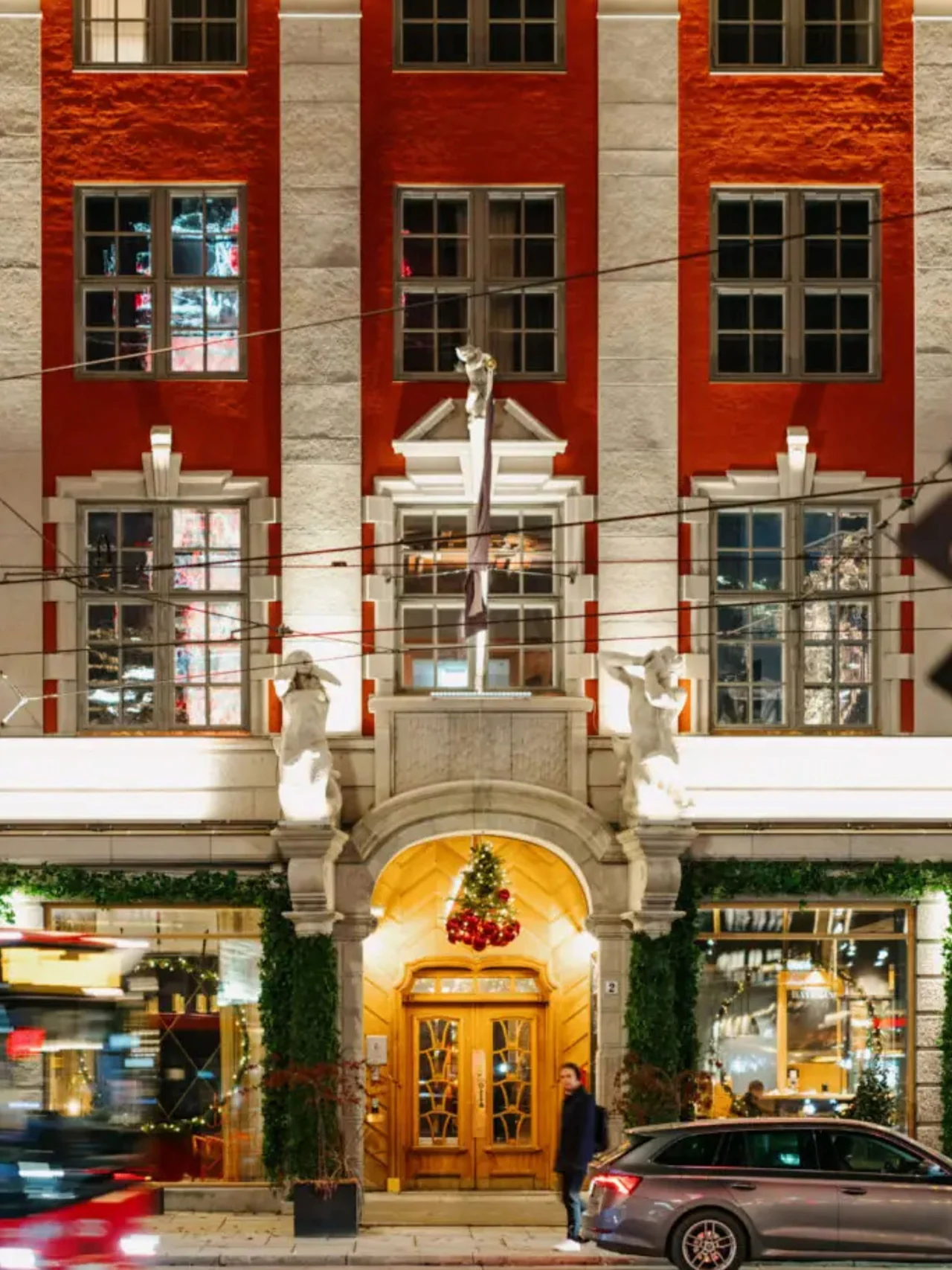En lysende rød-hvit bygning har statuer og juledekorasjoner over en buet tredør. En person går forbi, og en bil og buss passerer i forgrunnen.