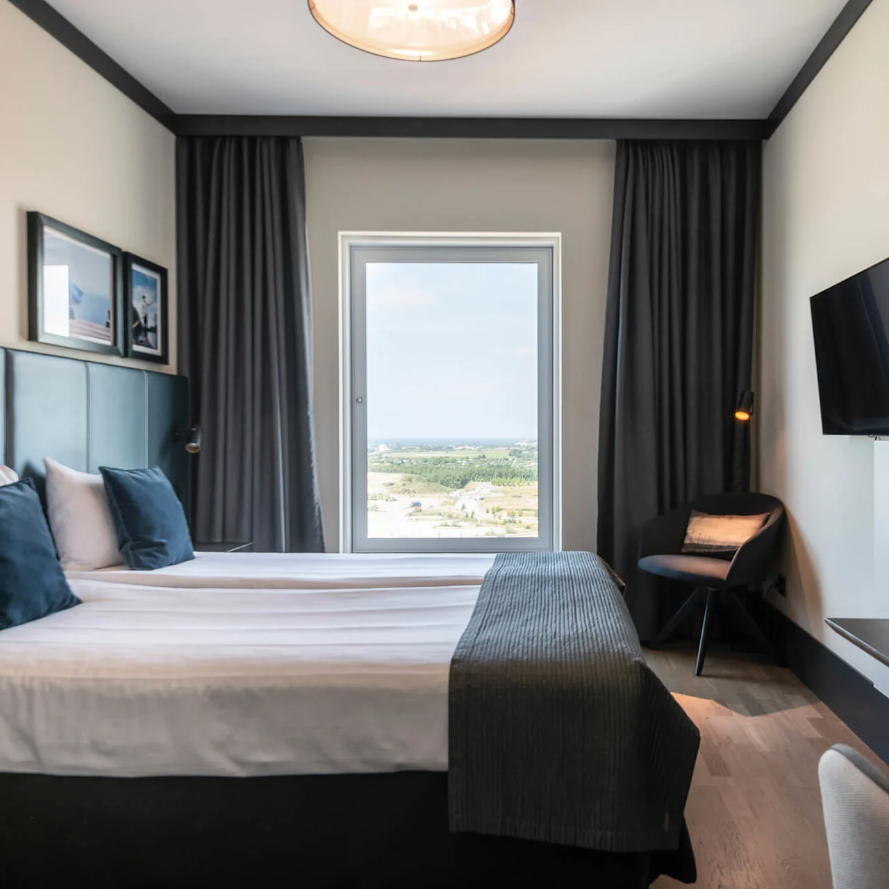 A neatly made bed with pillows rests beside a large window, revealing a scenic landscape. A wall-mounted TV faces a nearby chair and desk in a modern room.