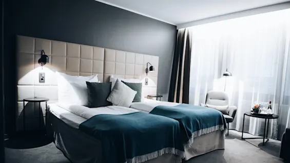 Two neatly made twin beds with blue throws and pillows sit in a modern, softly lit hotel room with a gray accent wall, beside a window with sheer curtains.