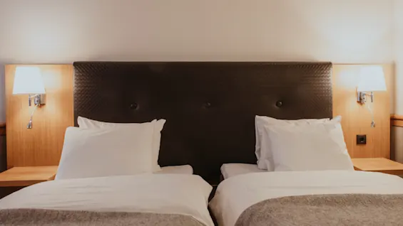 Two comfortable single beds with white linens and grey throws in a hotel room, illuminated by wall lamps.