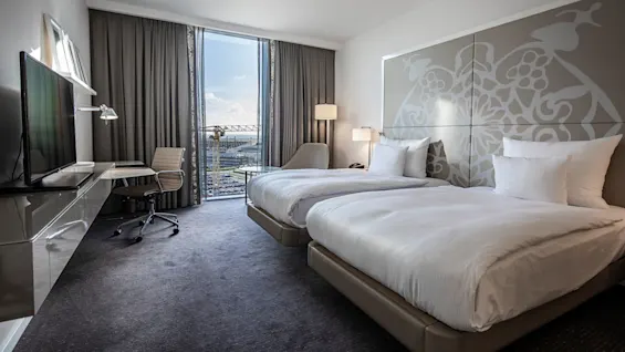 Två prydligt bäddade sängar med vita lakan står i ett elegant inrett hotellrum, upplyst av naturligt ljus från ett stort fönster med utsikt över stadssilhuetten.