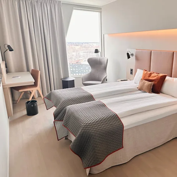 Ett prydligt hotellrum med en fint bäddad dubbelsäng och gråa och orange kuddar, samt ett arbetsbord, stol och fåtölj vid ett fönster. Rökning förbjuden-skylt finns.