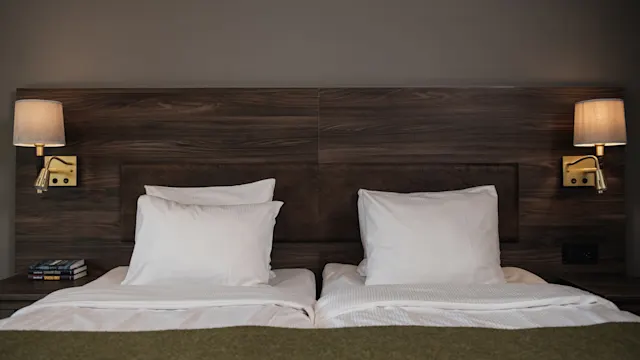 A neatly made double bed with white pillows and sheets, flanked by wall-mounted lamps and a wooden headboard; books rest on the left side table.