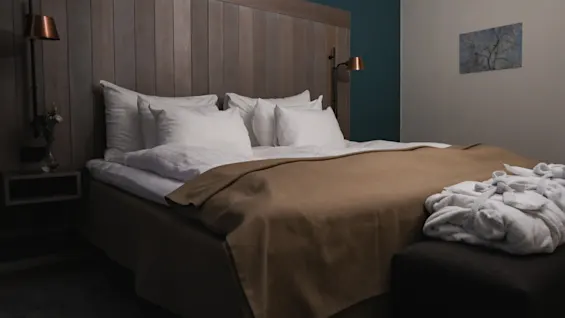 A neatly made bed with white pillows and a brown blanket sits beneath a wooden headboard. Two robes lie on a bench, in a softly lit room with teal and cream walls.