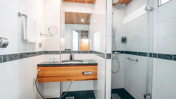 A modern bathroom features a wooden vanity with a sink, set against white tiled walls. A glass-enclosed shower is adjacent, with overhead lighting providing a bright, clean atmosphere.