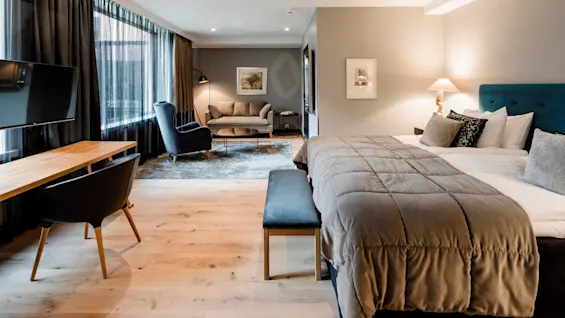Et moderne hotellrom har en stor seng med polstret hodegjerde, en koselig sittegruppe med sofa og lenestol, og et skrivebord i tre med TV, alt på tregulv.