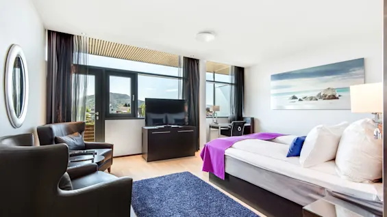 Et moderne hotellrom med en pent oppredd seng dekket med lilla detaljer, vendt mot en stor flatskjerm-TV. To lenestoler og et skrivebord er nær vinduet med fjellutsikt.