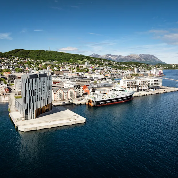 Quality Hotel Harstad, en moderne bygning på en brygge i en livlig byhavn med et stort skip fortøyd, omgitt av åser og fjell.