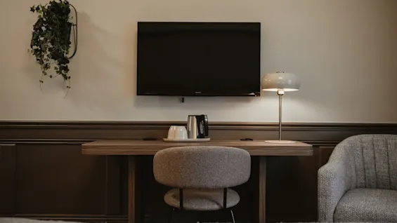 Et koselig hotellrom med skrivebord, stol, TV og en varm lampe, perfekt for avslapning eller en rask arbeidsøkt.