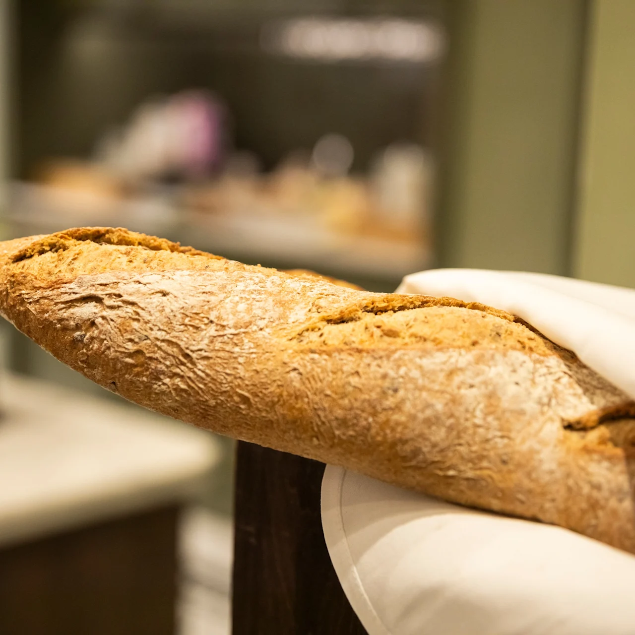 Loaf of rustic bread wrapped partially in a white cloth, resting on a kitchen counter with blurred background of kitchen items.