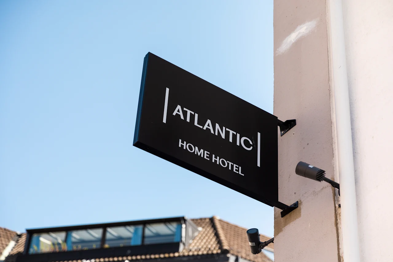 Et sort skilt med hvid tekst, der læser ATLANTIC HOME HOTEL, fastgjort til en bygning mod en klar blå himmel.