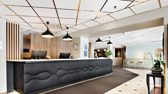 Vastaanottotiski, mustalla tasolla, varustettu tietokoneilla, ripustettujen lamppujen alla; puupaneelien ja koristekasvien ympäröimänä, viereisessä istuma-alueessa tuoleja ja pöytä.