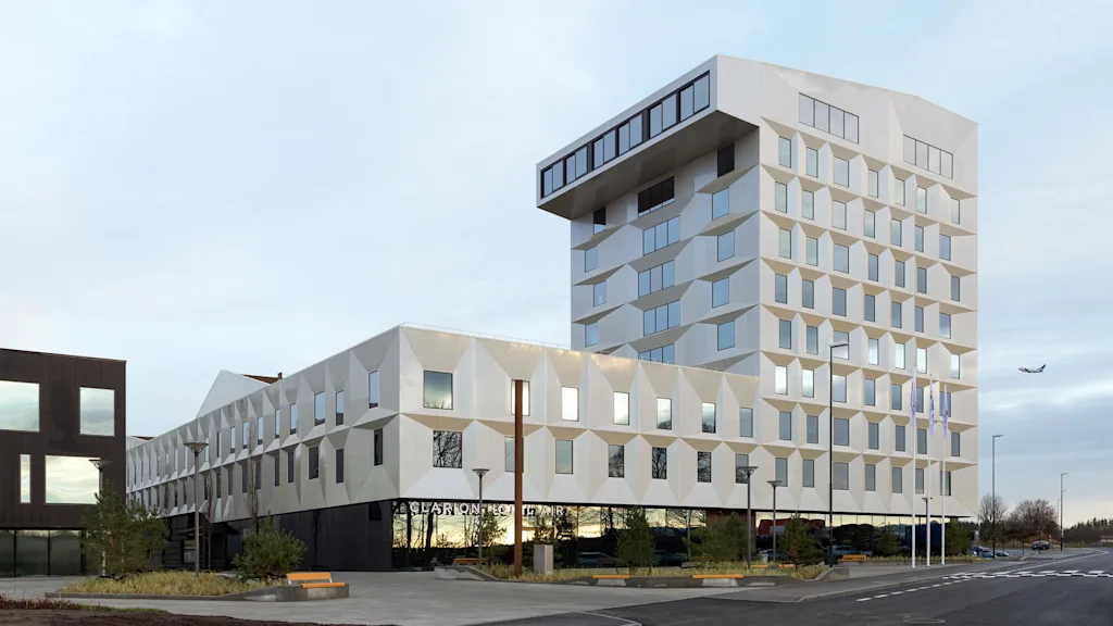 En modern, geometriskt designad byggnad med vinklade fönster och vit fasad står högt. Nedan finns texten "CLARION HOTEL AIR." I närheten flyger ett flygplan i en delvis molnig himmel.