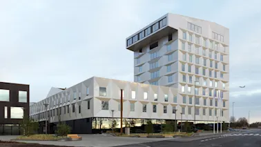En moderne, geometrisk designet bygning med vinklede vinduer og hvit fasade står høyt. Nedenfor står teksten "CLARION HOTEL AIR." I nærheten flyr et fly i en delvis skyet himmel.