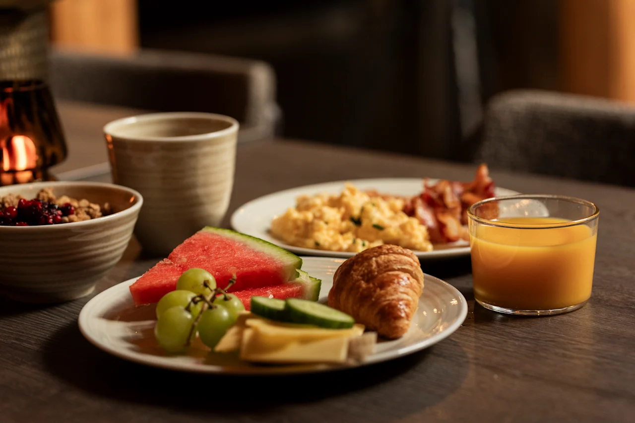 Et dejligt morgenmadsbord på et træbord, med frisk frugt, en croissant, røræg, bacon og juice.