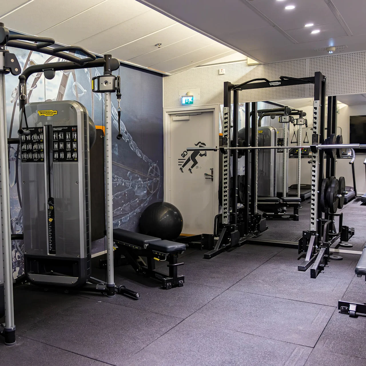 A gym features various workout machines and benches, with weights and a stability ball. A clock is on the wall next to a poster of a running figure. 