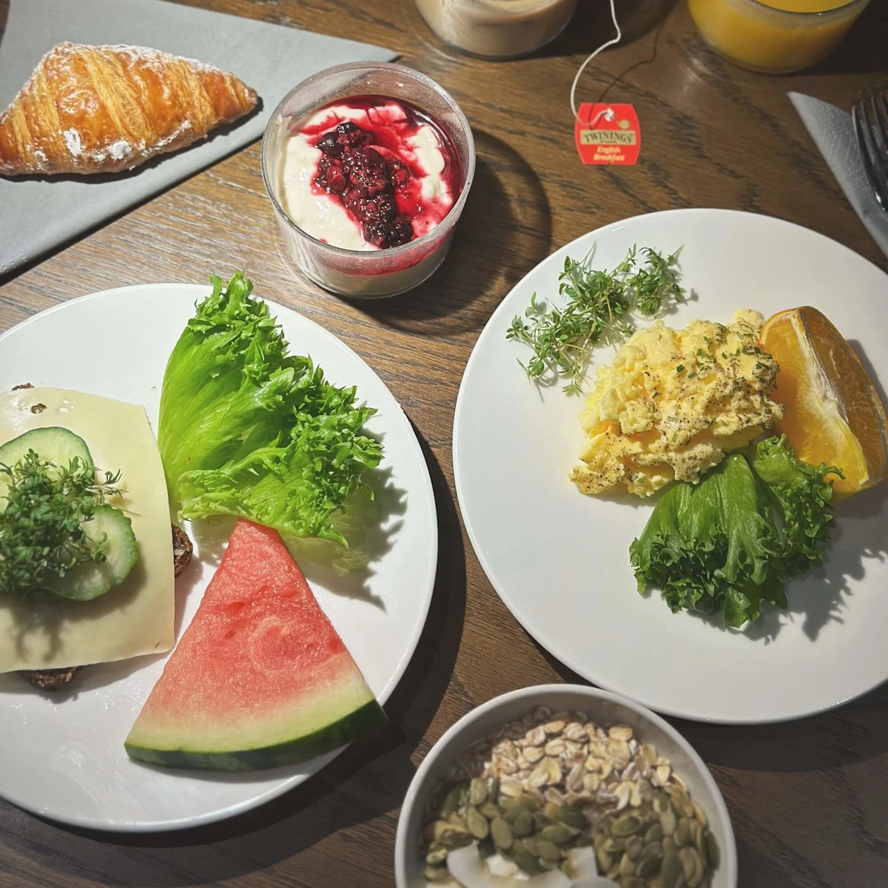 A croissant, plate with scrambled eggs, toast with cheese, vegetables, and fruit. Yogurt with berry topping, muesli bowl, cups of tea ("Twinings English Breakfast") and orange juice on a wooden table.
