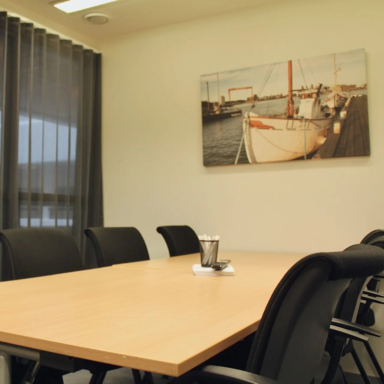 A rectangular conference table is surrounded by black chairs in a room with large windows and sheer curtains. A framed picture of a docked sailboat hangs on the wall.