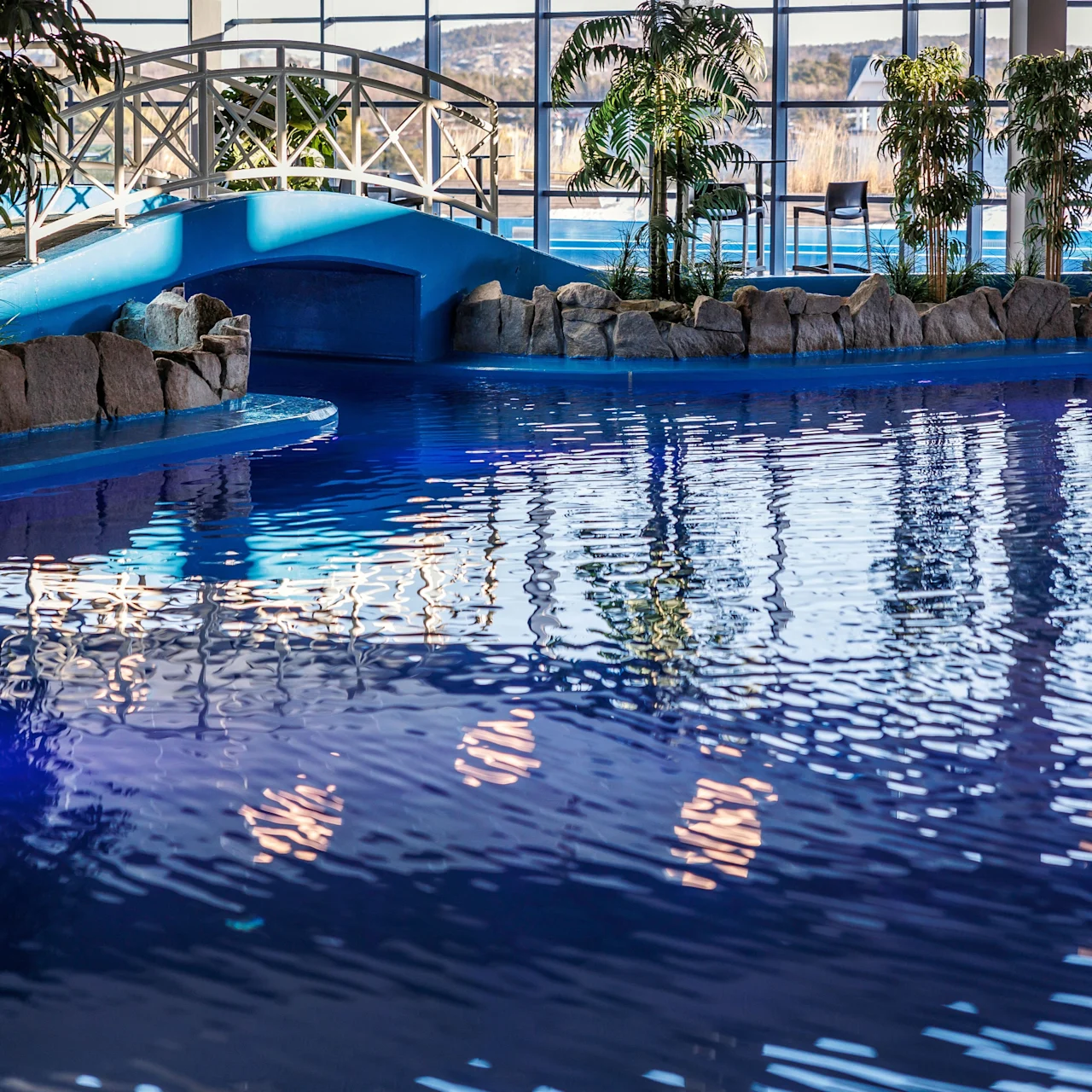 A swimming pool reflects soft lighting; surrounded by tropical plants and a small arched bridge, creating a serene atmosphere indoors with large windows offering a view of a distant landscape.