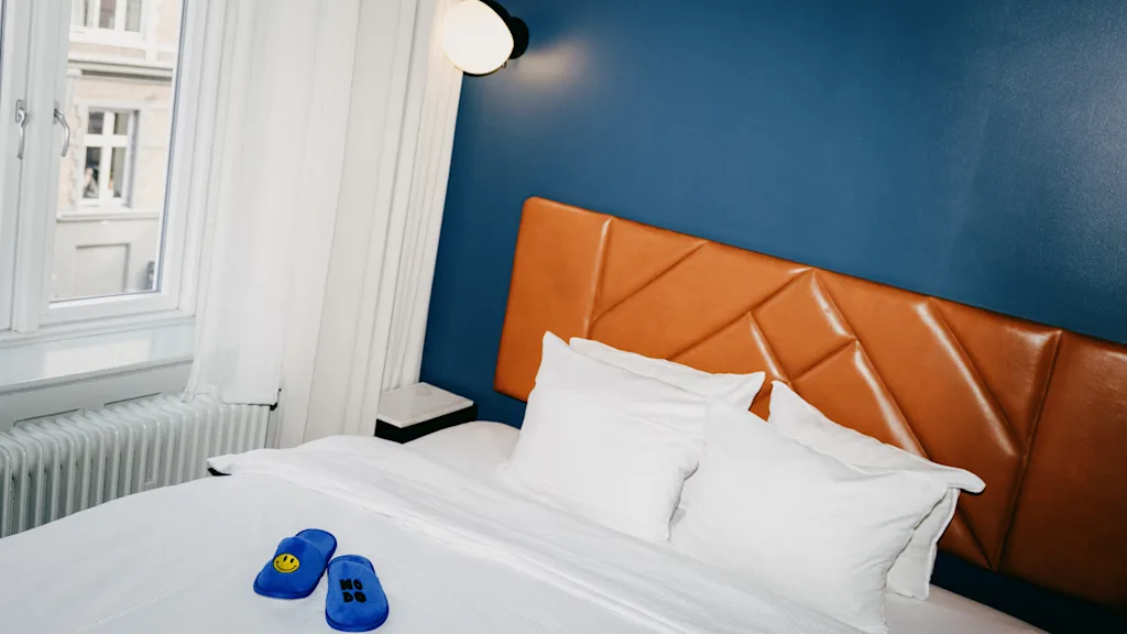 Moderni hotellihuone, jossa on sininen seinä, nahkainen sängynpääty ja siniset tossut sängyllä.