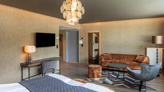 Ett hotellrum har en säng med ett grått quiltat täcke, en mysig sittgrupp med orange möbler och en platt-TV på väggen. Modern inredning kompletterar den varma belysningen.