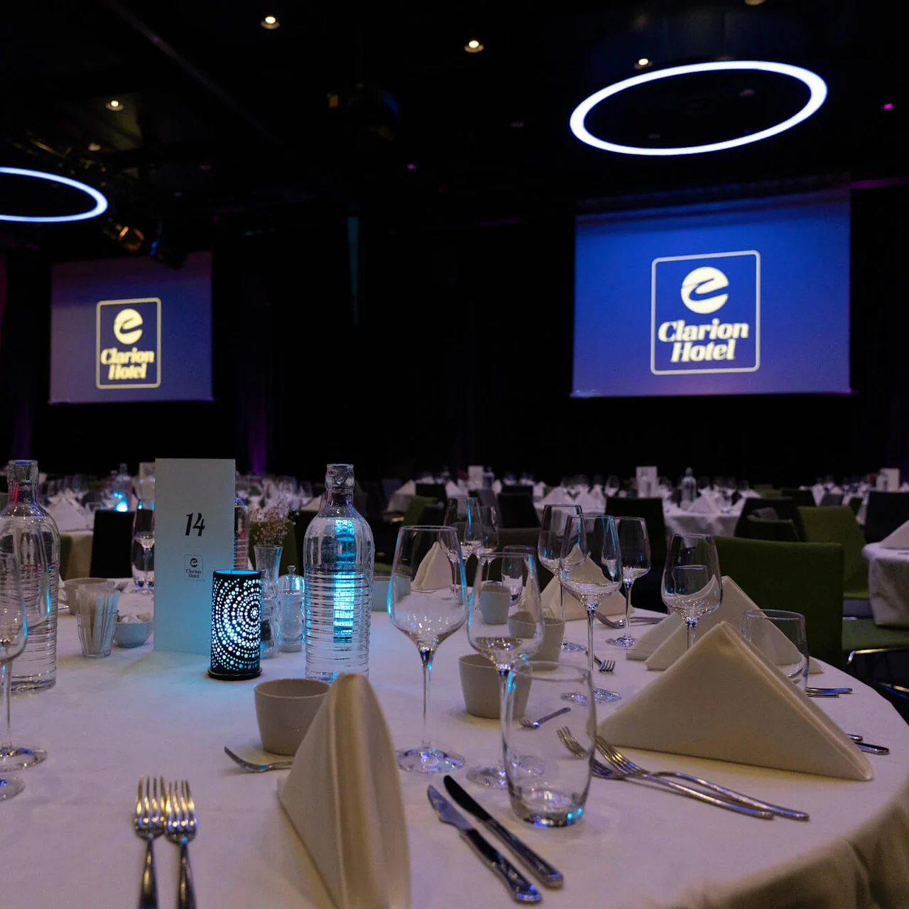 Et rundt banketbord er elegant dækket med glas og servietter i en svagt oplyst eventhal. Skærme viser "Clarion Hotel"-logoen. Bordkortet lyder "14."