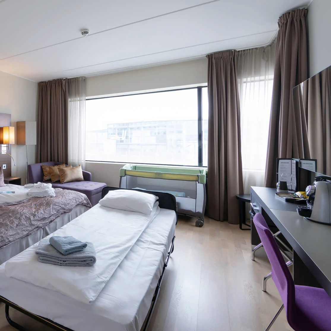 A neatly arranged hotel room features two beds with crisp white linens, a purple chair by a sleek desk, and a comfortable sofa near a large window. Folded towels on beds.