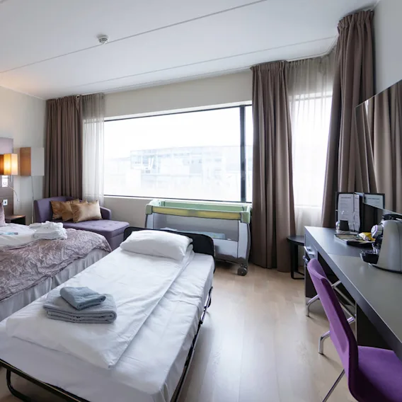 Et ryddig hotellrom med to senger med hvitt sengetøy, en lilla stol ved et stilig skrivebord og en komfortabel sofa nær et stort vindu. Brettede håndklær på sengene.