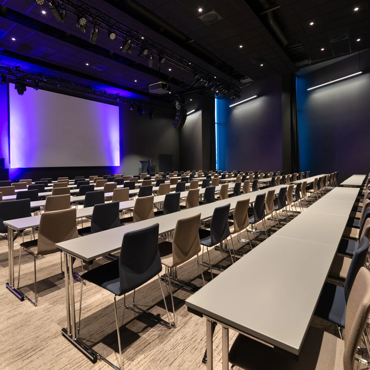 Rader av seter vendt mot en stor projeksjonsskjerm i et konferanserom med nøytrale veggfarger og blå belysning.