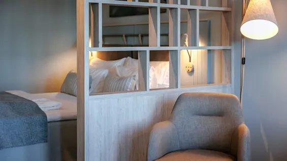 A beige armchair sits beside a wooden partition with open sections. Behind it, a neatly made bed with striped cushions is illuminated by a wall lamp, creating a cozy atmosphere.