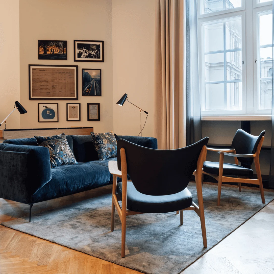 Blue velvet sofa with patterned cushions sits on a gray rug, facing two wooden chairs near large window. Framed art decorates beige walls; lamps add soft lighting.