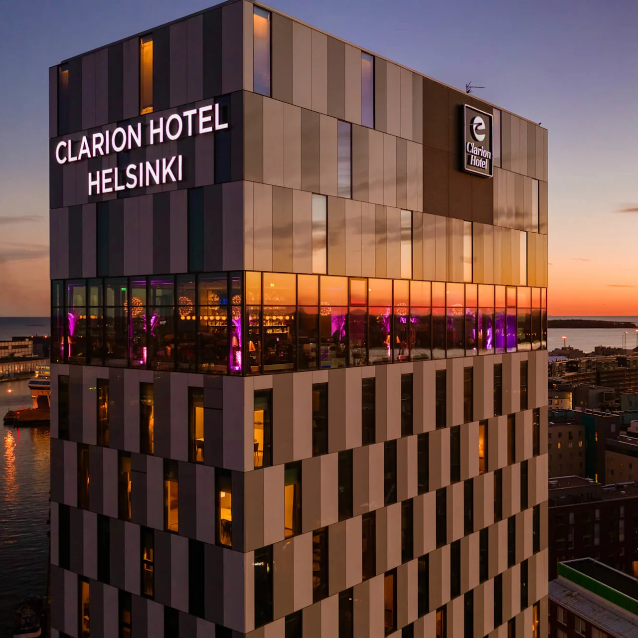 Clarion Hotel Helsinki tårner sig over en havn ved solnedgang, med reflekterende vinduer oplyst af lilla lys indendørs. Den omkringliggende by og vand skaber en rolig aftenstemning.