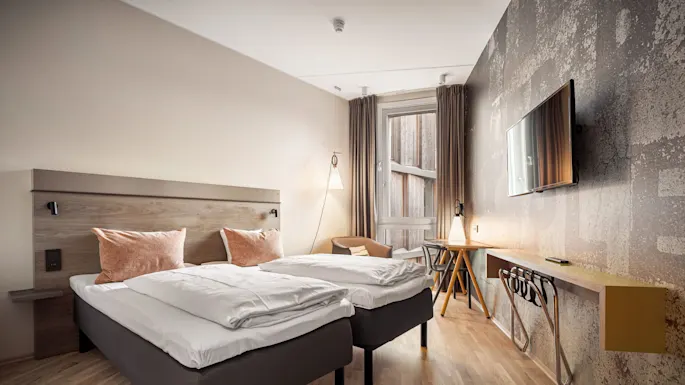 Two neatly made twin beds stand in a modern hotel room, beside a light-filled window, with wooden flooring and a wall-mounted television opposite.