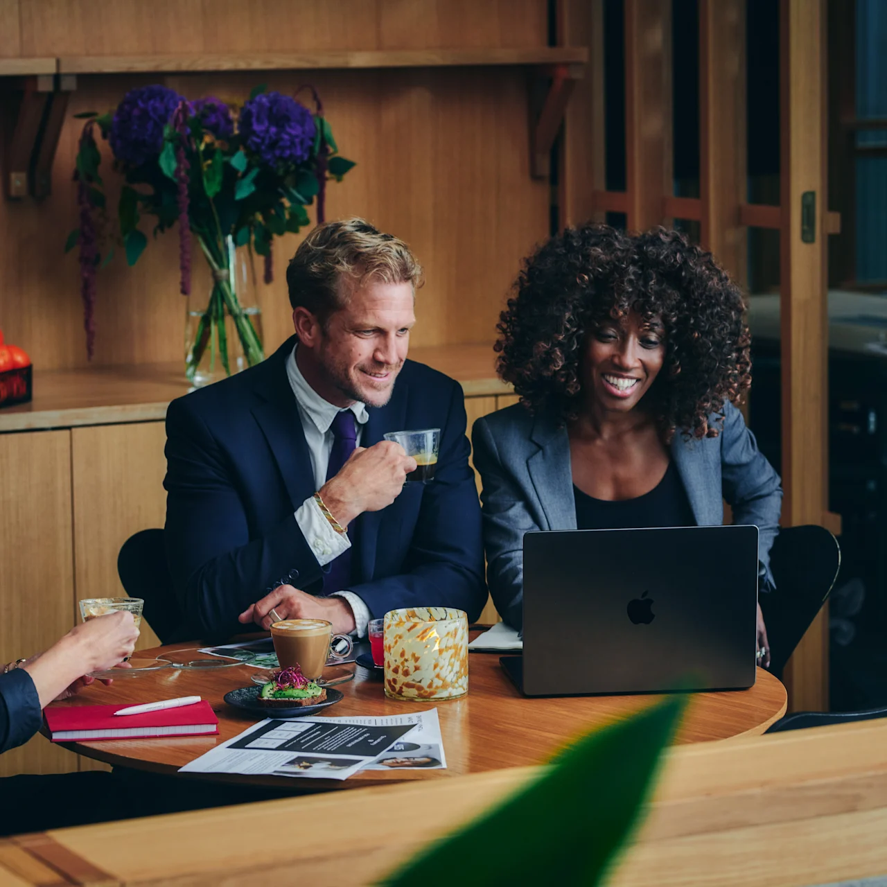 En bärbar dator står öppen på ett runt bord, omgiven av tre leende människor som dricker kaffe. Bakgrunden innehåller trähyllor, en vas med blommor och en skål med apelsiner. Ingen text finns.