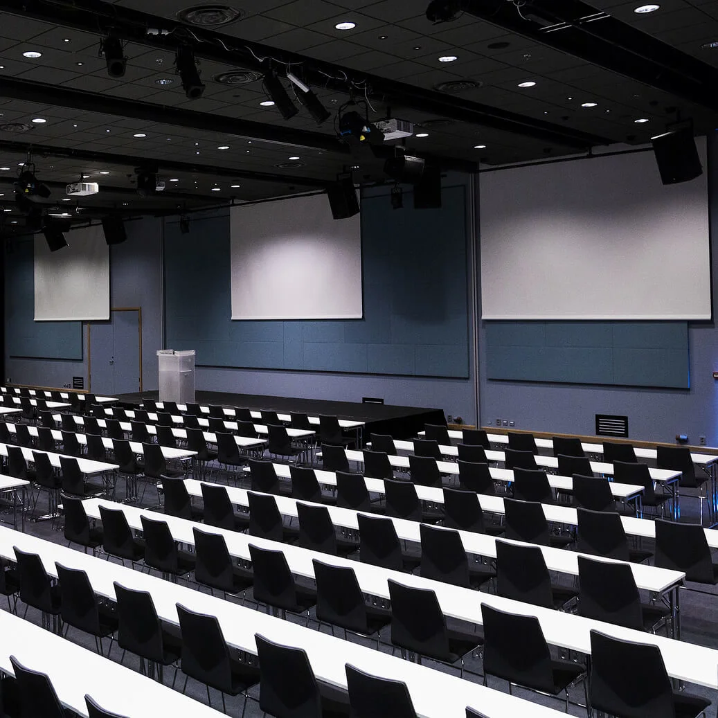 Empty auditorium with rows of white tables and black chairs, facing multiple large projection screens on a stage, under dim ceiling lights.