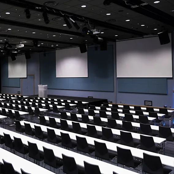 Tomt auditorium med rader av hvite bord og svarte stoler, vendt mot flere store projektorskjermene på en scene, under svake taklys.