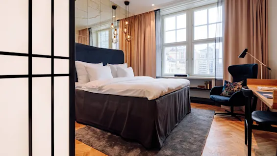 Bed with white linens, positioned against a mirrored wall, surrounded by hanging lights and large window, located in a modern room with wood flooring, chair, and desk.
