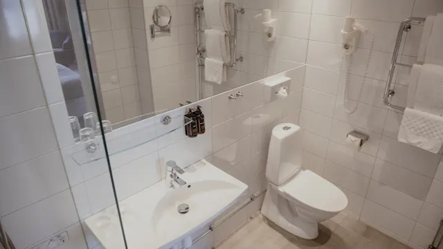 En vit keramisk toalett står i ett kaklat badrum; bredvid ett handfat med spegel. På väggen finns handdukshållare och en hårtork. Handdukar är prydligt upphängda.