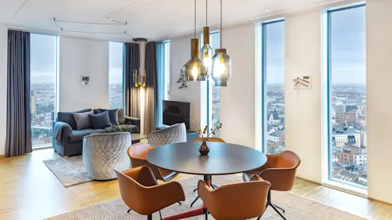 Modernt matbord med bruna stolar står under hänglampor i ett rymligt vardagsrum. Stora fönster ger utsikt över stadssilhuetten, med en grå soffa och TV som kompletterar scenen.