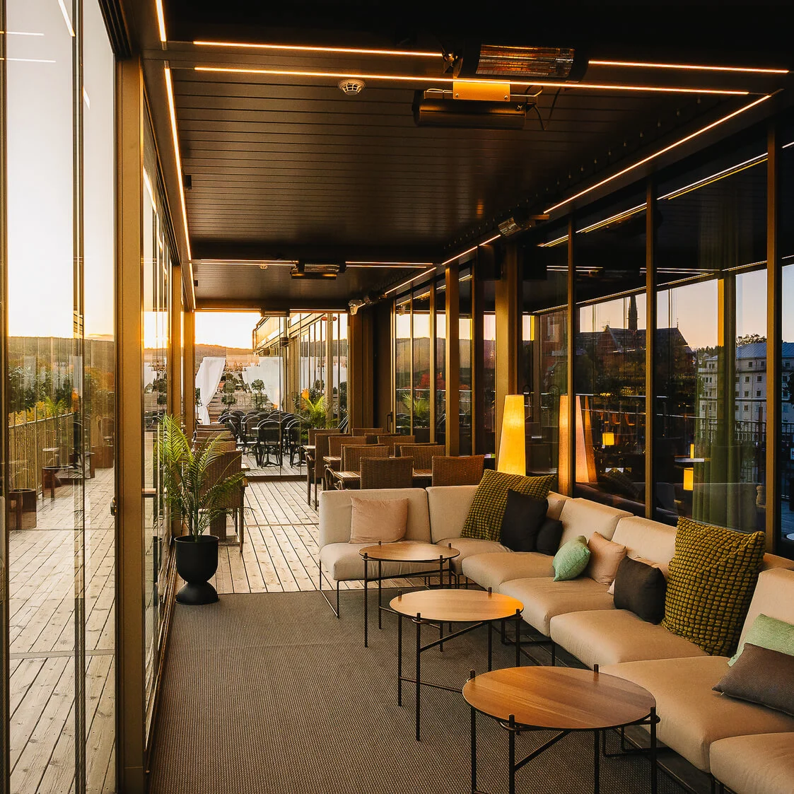 En moderne terrasse med hyggelige siddepladser har neutrale sofaer og små borde. Udsigt over et bylandskab med en fremtrædende kirke og træer, indrammet af store glasvinduer.