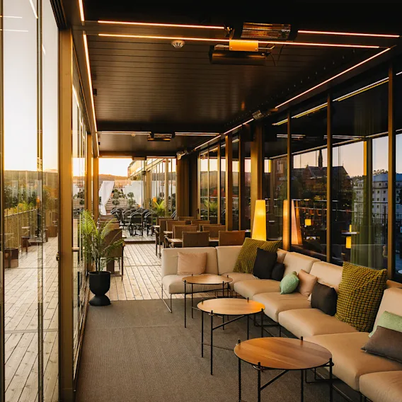 A modern terrace with cozy seating features neutral couches and small tables. It overlooks a scenic cityscape with a prominent church and trees, framed by large glass windows.