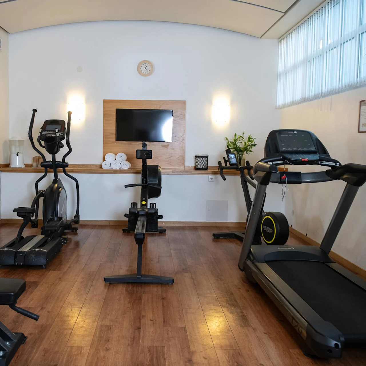 A compact gym features a treadmill, elliptical, and rowing machine on a wooden floor. A wall-mounted TV, clock, and row of neatly rolled towels enhance the well-lit, tidy space.