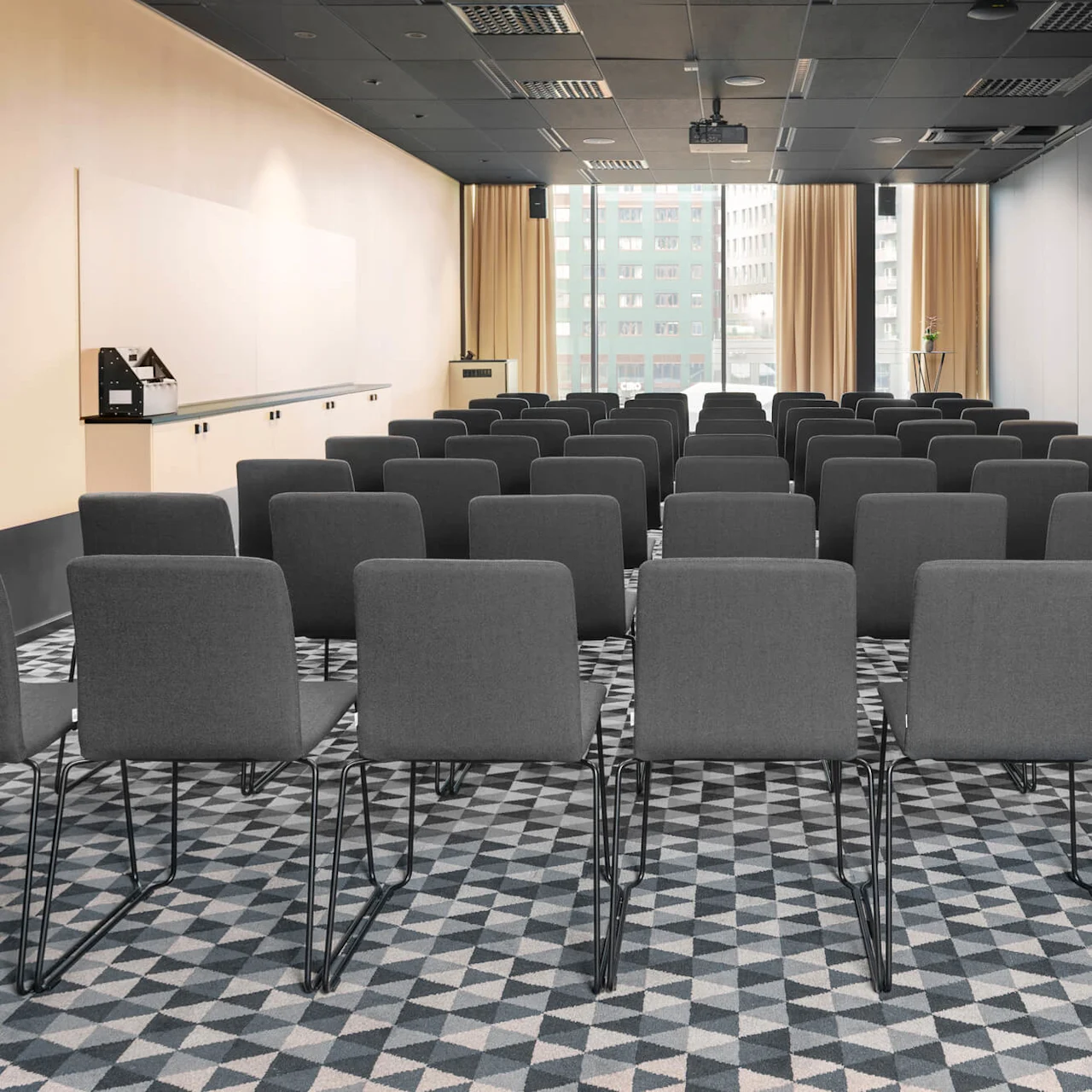 Rader med grå stolar står vända mot en tom vägg i ett modernt konferensrum med mönstrad matta och fönster från golv till tak, upplyst av naturligt ljus.
