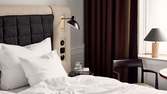 Bed with white pillows and sheets, illuminated by a black wall lamp. Nearby, a glass of water and book rest on a small table. Dark curtains frame a window with another lamp.