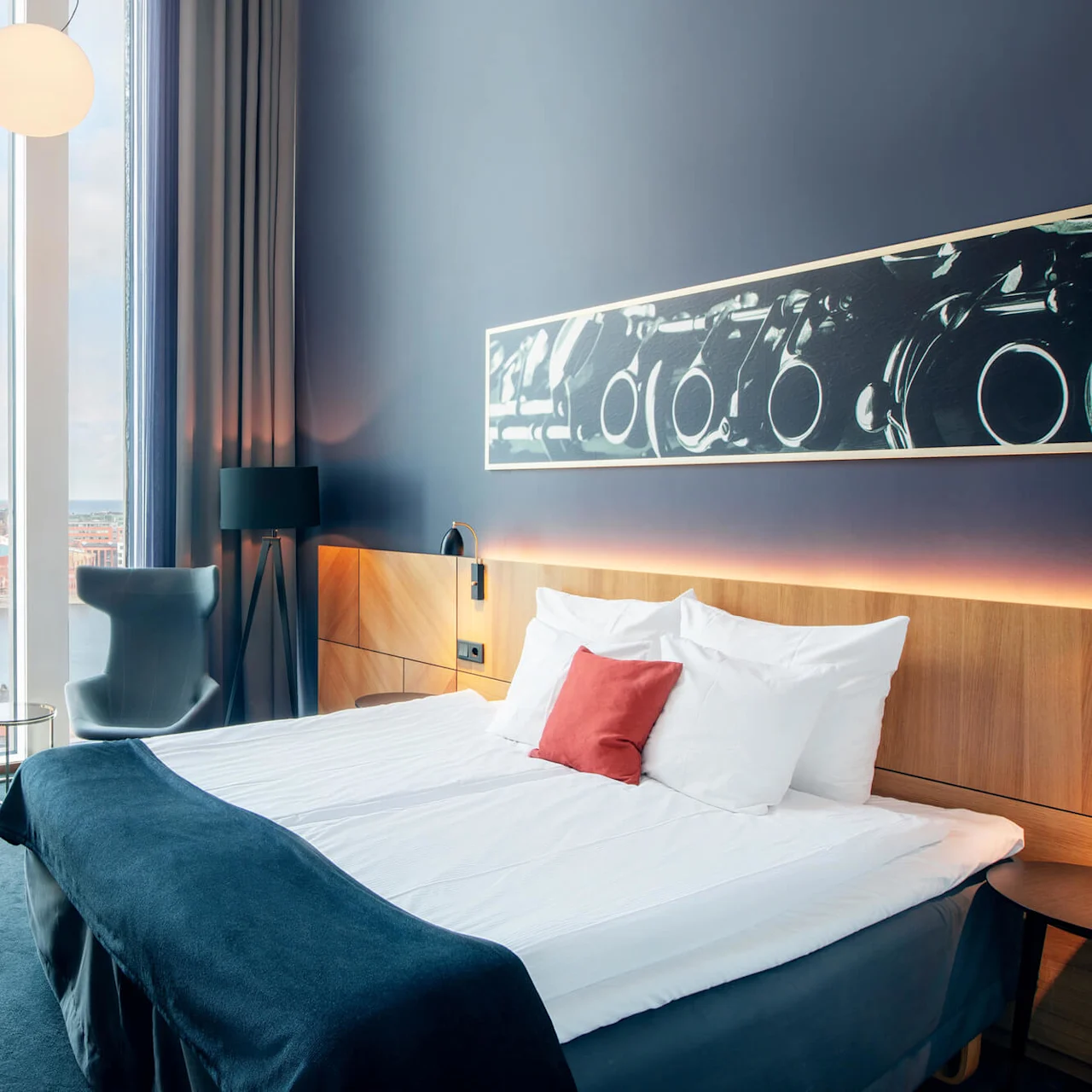 En stor seng med hvitt sengetøy og en rød pute står i et moderne hotellrom med gulv-til-tak-vinduer og byutsikt. Et skrivebord og en stol er i nærheten.