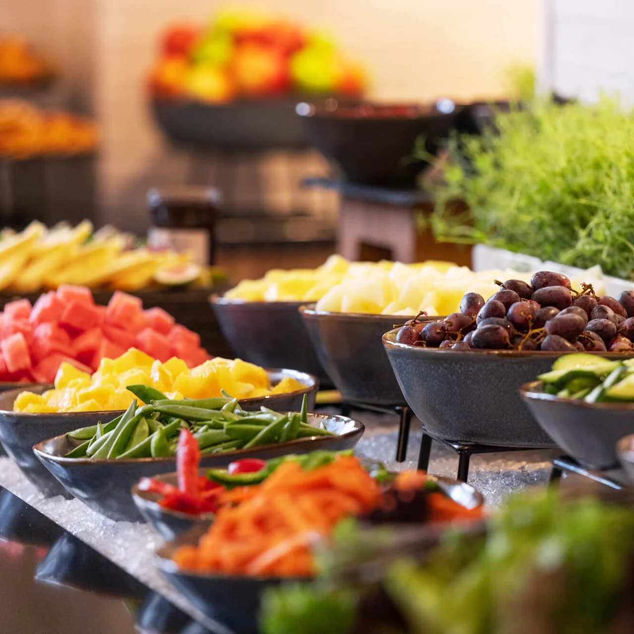 Skåle fyldt med blandet frugt og grøntsager, herunder druer, agurker og tomater, er pænt arrangeret på en buffet. Usædvanlig grønt i baggrunden tilføjer friskhed.