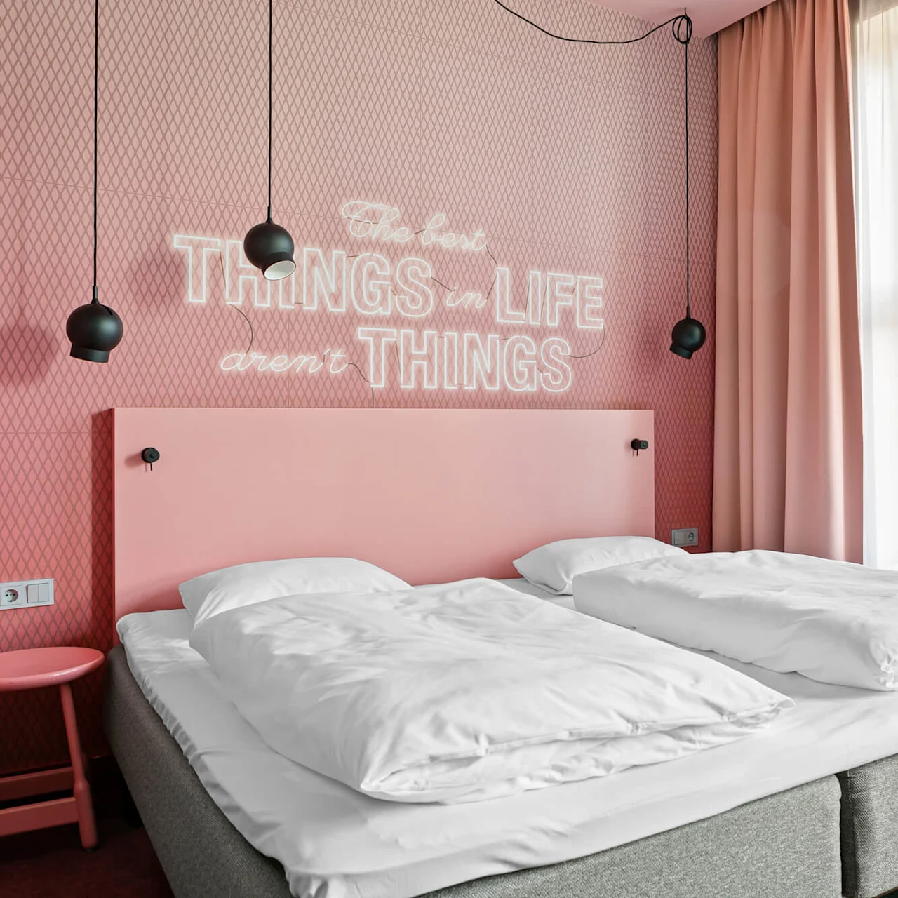 Seng med hvitt sengetøy ved siden av rosa vegg med glødende tekst "The best things in life aren't things," med hengende svarte lamper, ved siden av vindu med rosa og hvite gardiner.